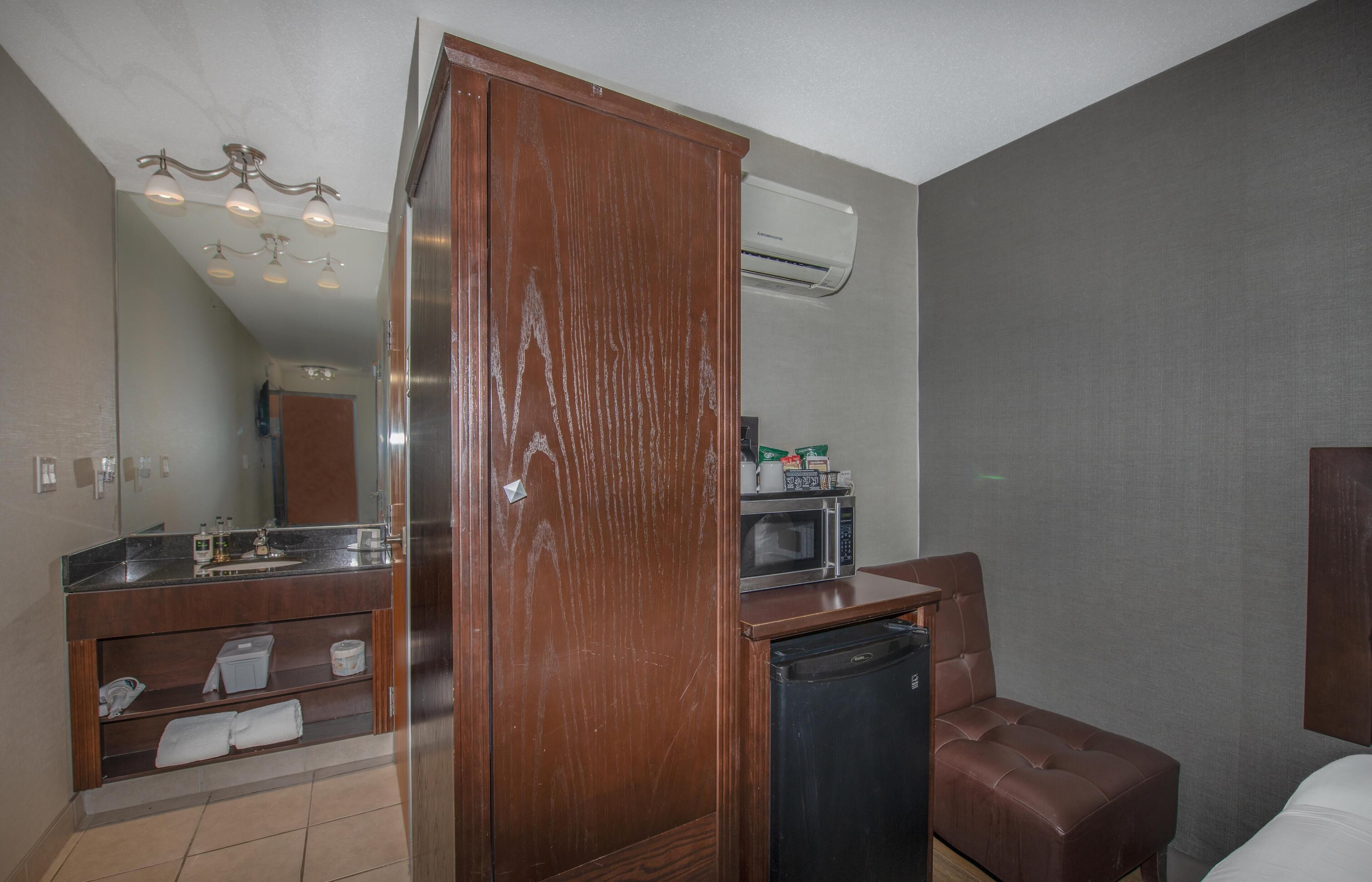 Quarto de hotel com guarda-roupa de madeira, micro-ondas, mini-geladeira, poltrona de couro marrom e uma cama.