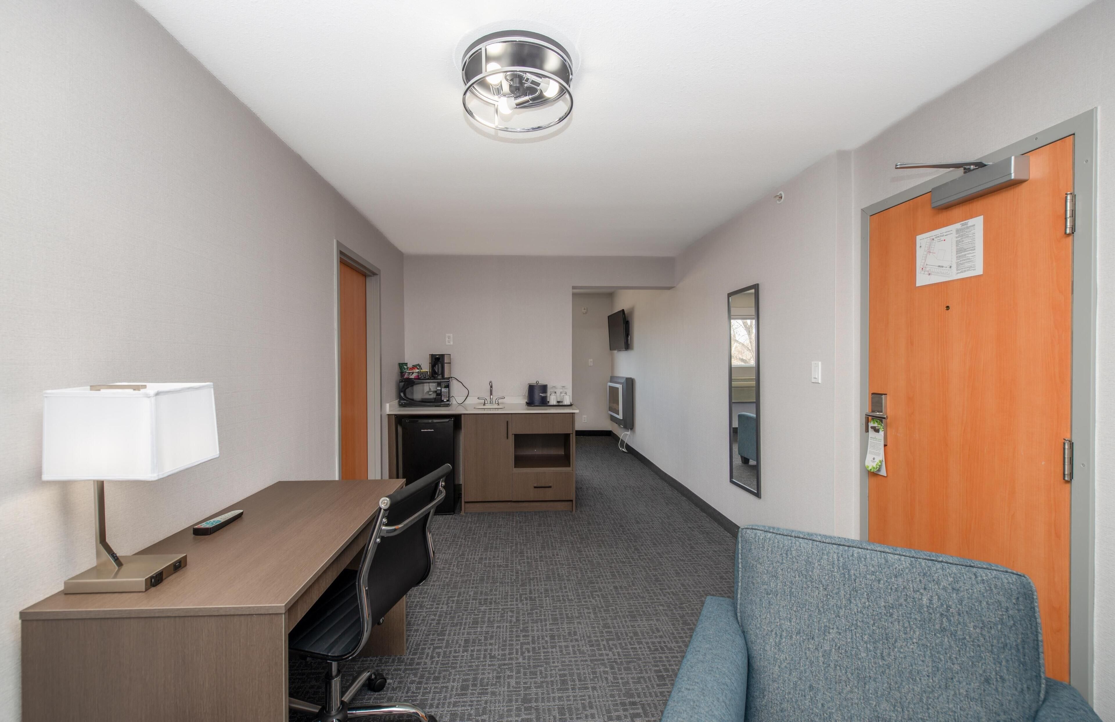 A hotel room with a desk, chair, sofa, kitchenette, and a door with a sign.