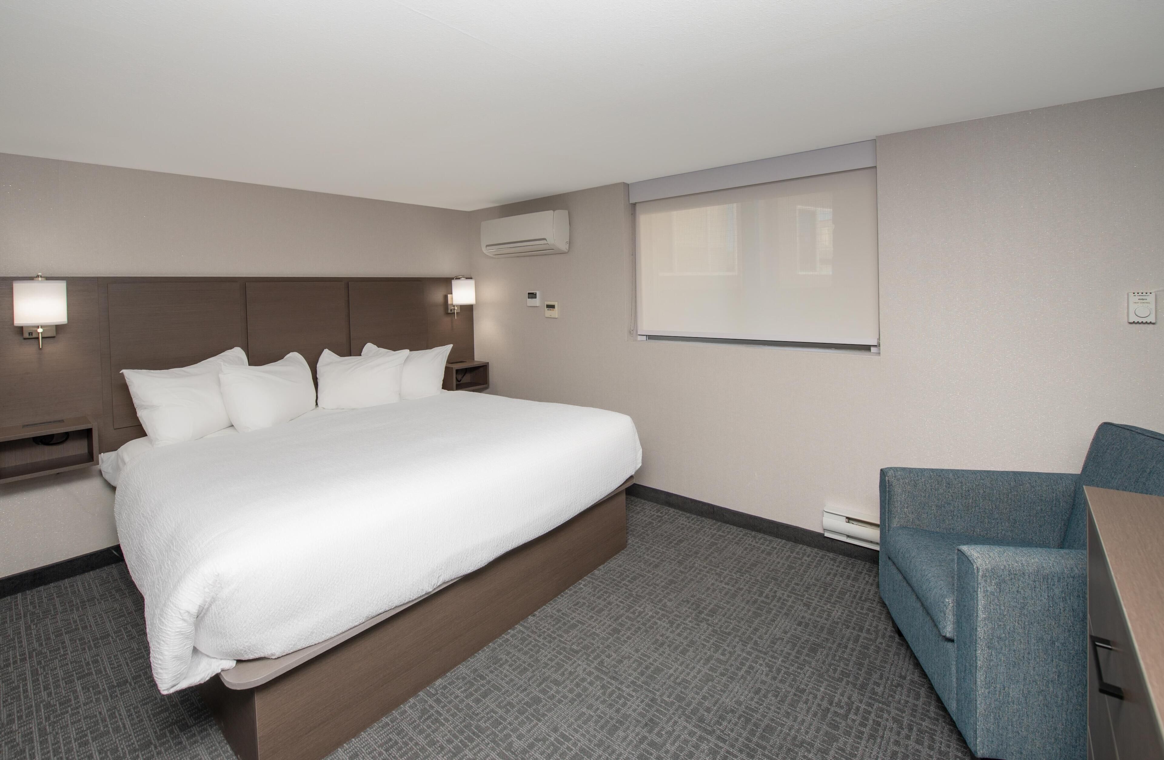A hotel room with a large bed, a blue armchair, a wall-mounted air conditioner, and a flat-screen TV.
