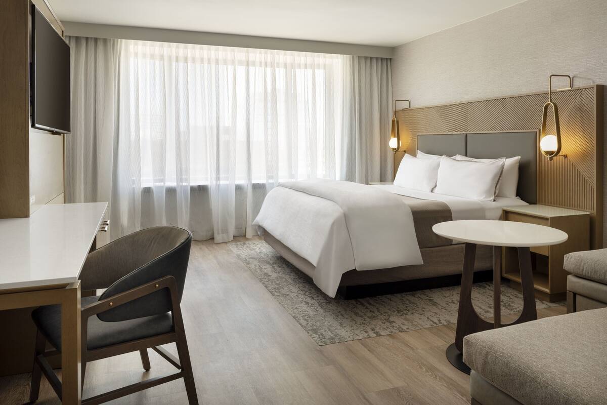 A hotel room with a bed, a desk, a chair, a small table, and a TV.