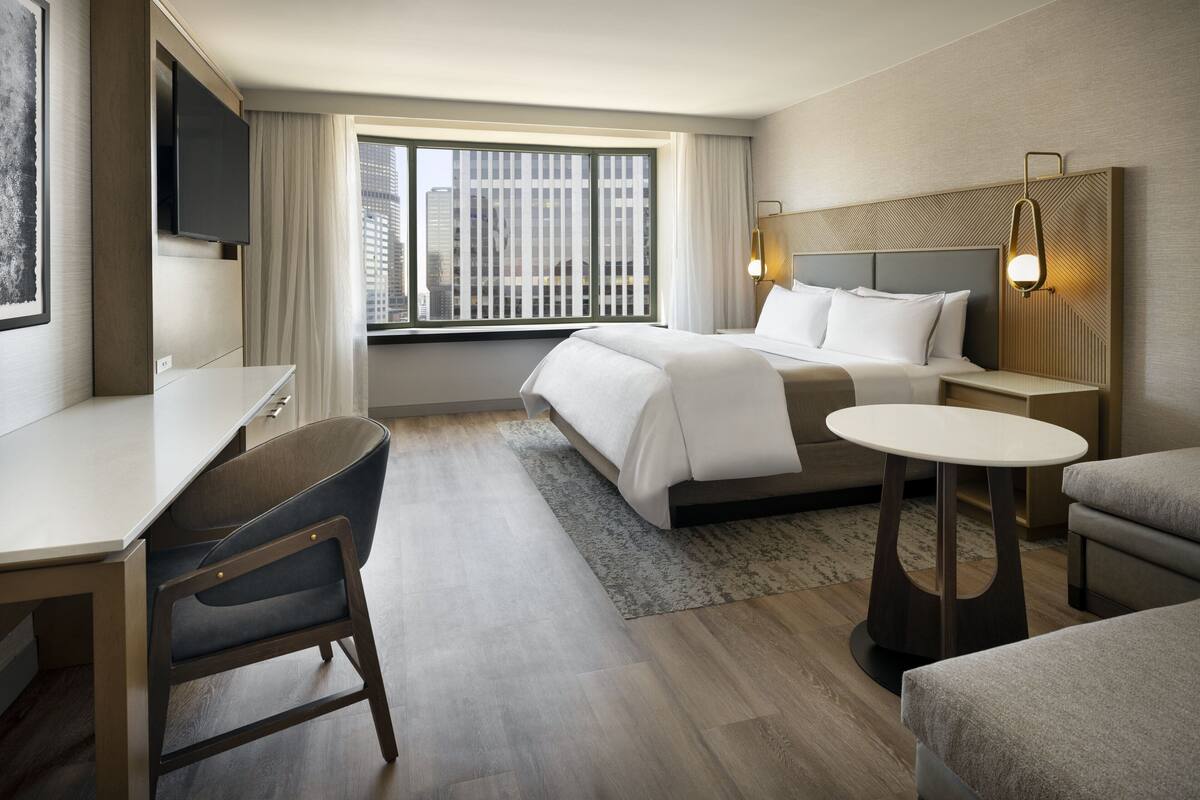 A modern hotel room with a bed, a desk, a chair, a small table, and a TV.