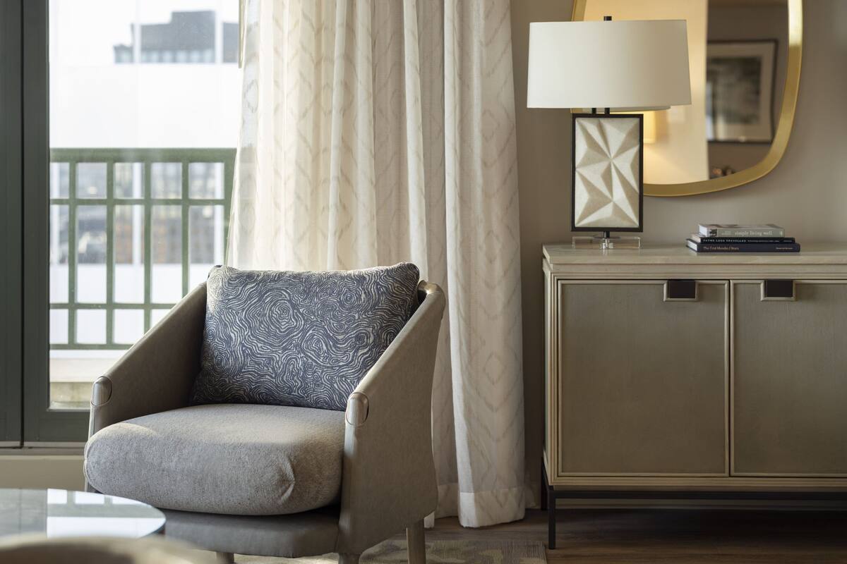 A modern hotel room with a sofa, a side table with a lamp, a framed picture, and a view of the city through a window with curtains.