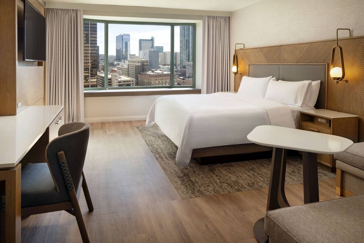 A hotel room with a large bed, a desk, a chair, and a view of the cityscape through a large window.