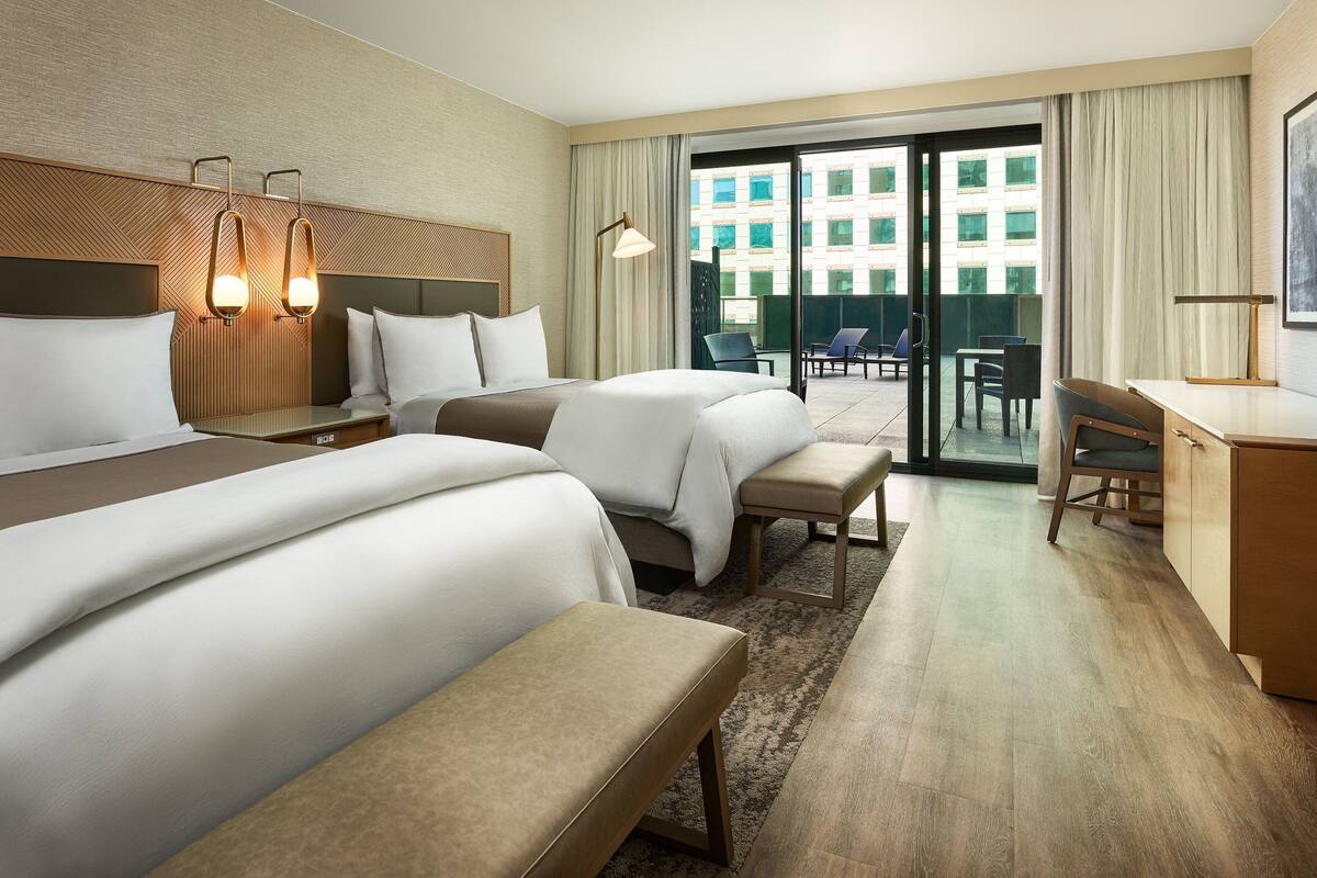 A hotel room with two beds, a desk, and a balcony with chairs.