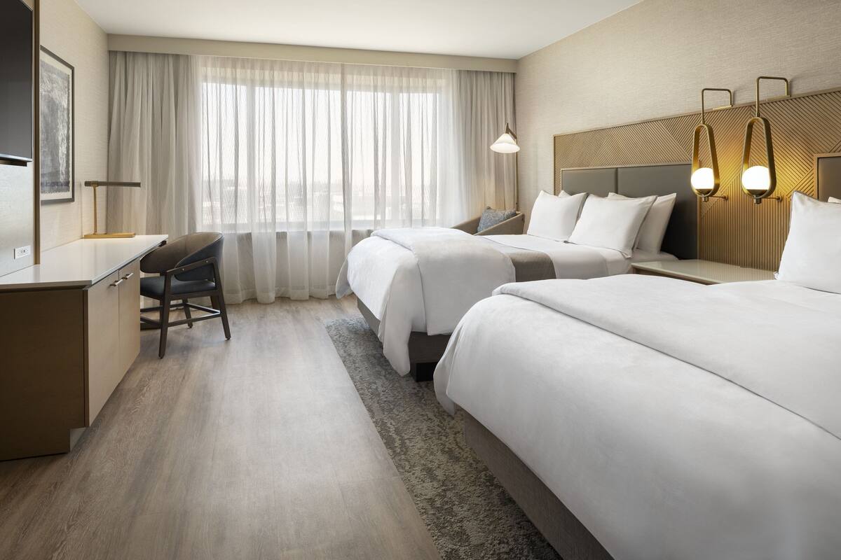 A hotel room with a large bed, a desk, a chair, and a window with sheer curtains.