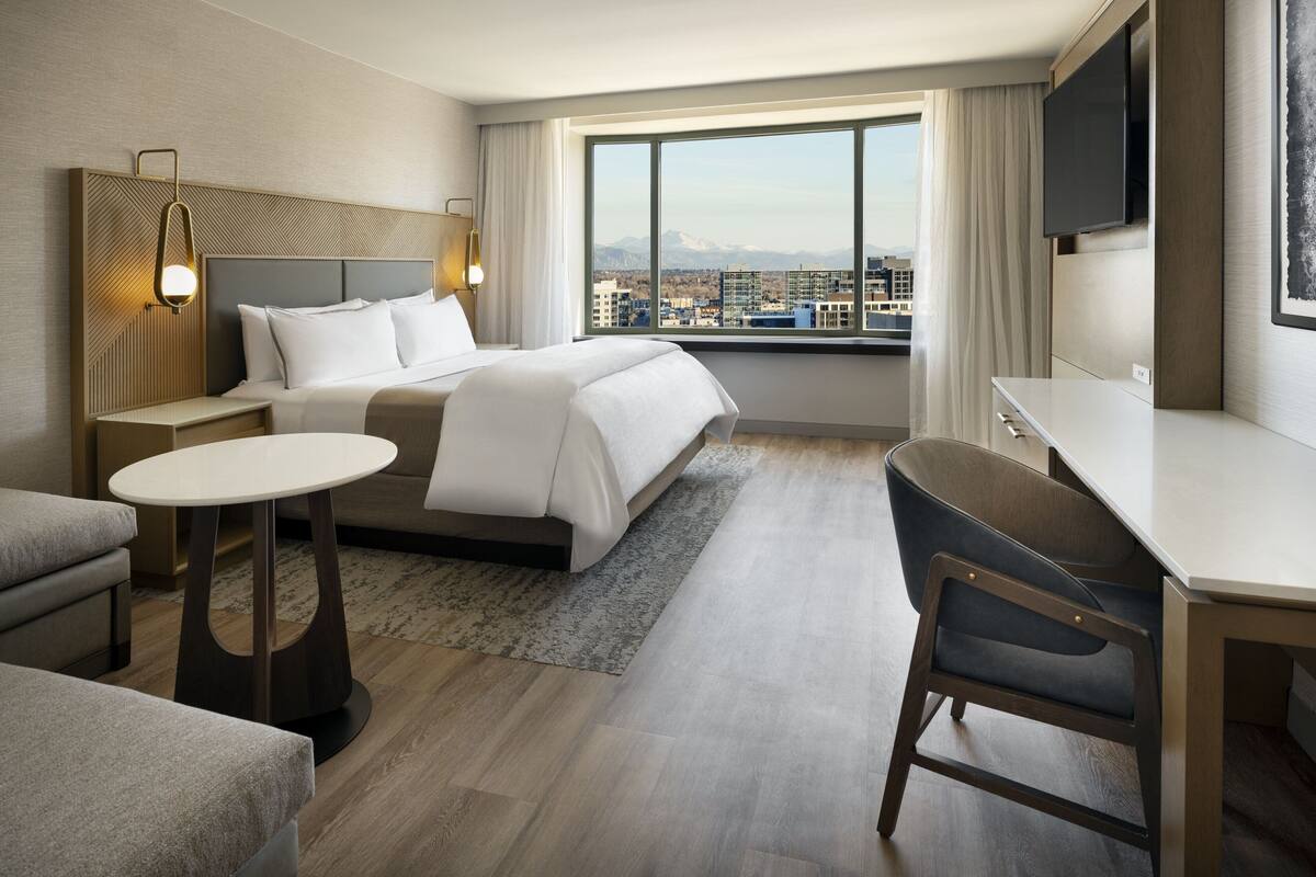 A modern hotel room with a large bed, a desk, a chair, and a view of the cityscape through large windows.