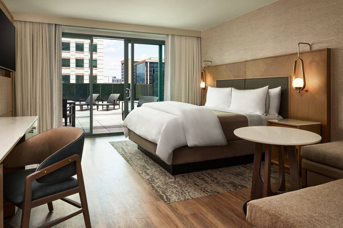 A hotel room with a bed, a desk, a chair, a small table, and a view of the outside.