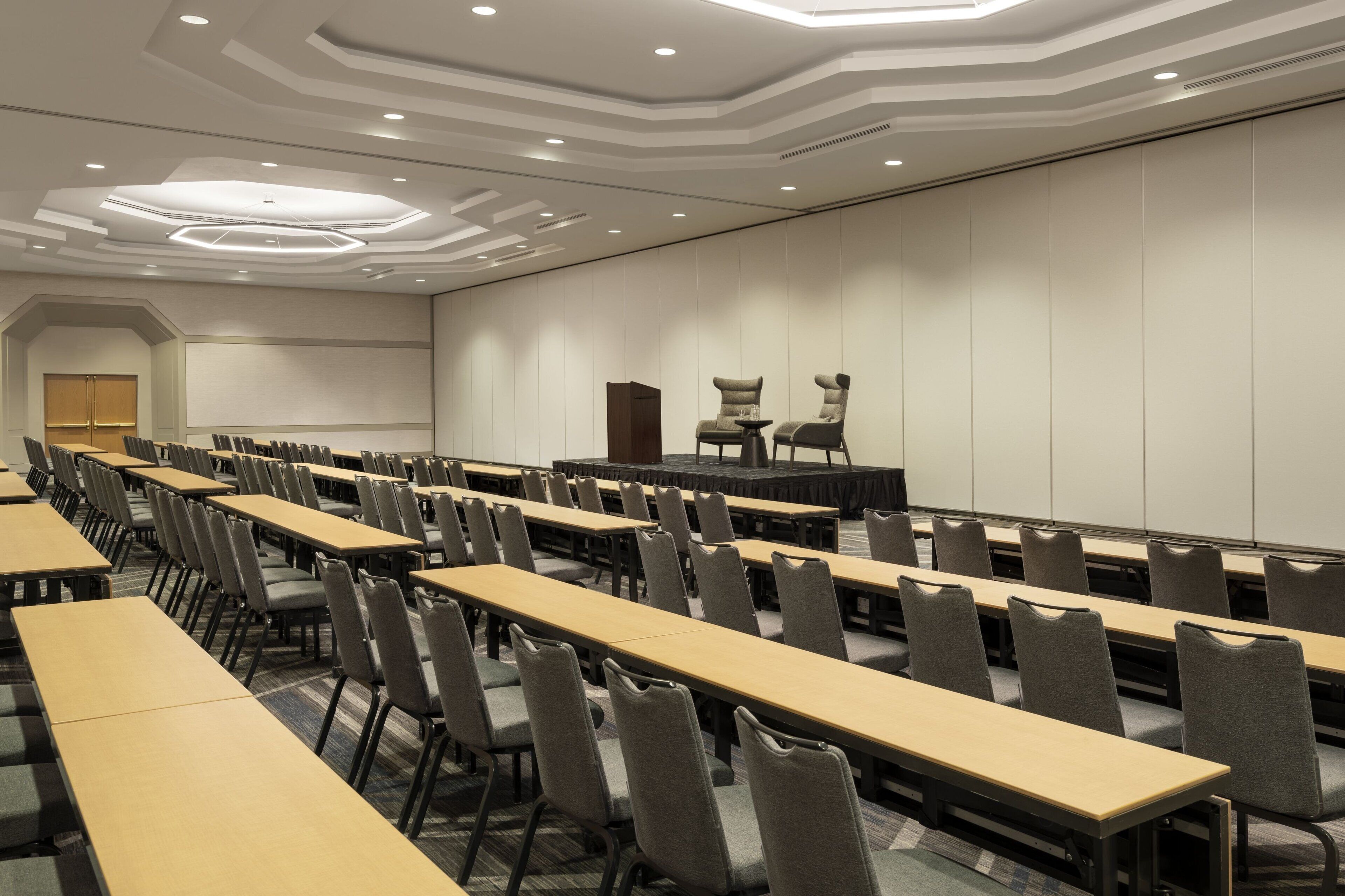 Une salle de conférence avec des rangées de tables et de chaises, une scène et un grand écran.