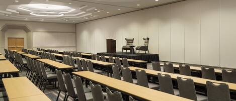 Une salle de conférence avec des rangées de tables et de chaises, une scène et un grand écran.
