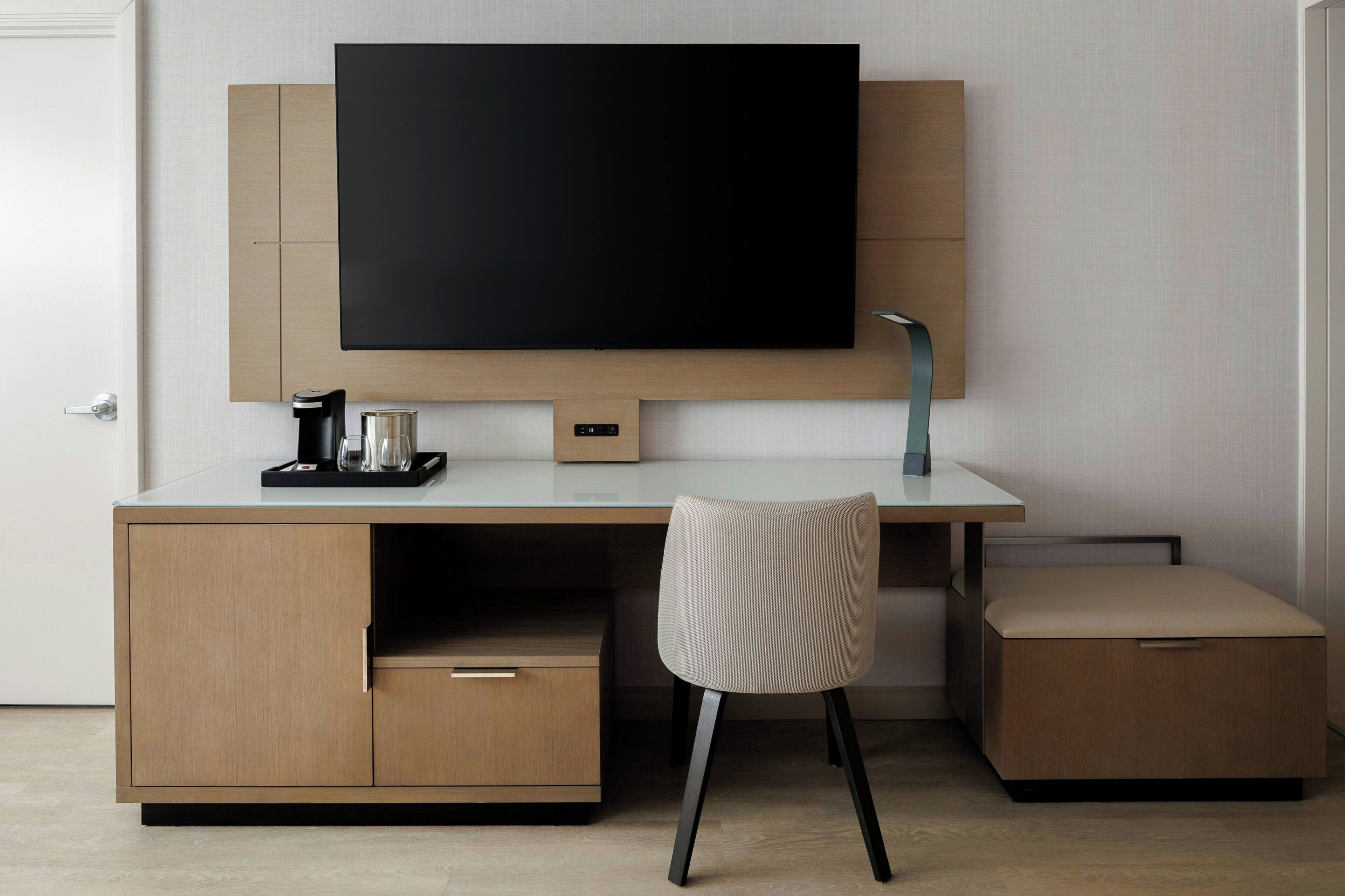 A modern hotel room with a large flat-screen TV mounted on a wooden cabinet, a small coffee maker, a glass of water, and a cushioned bench.