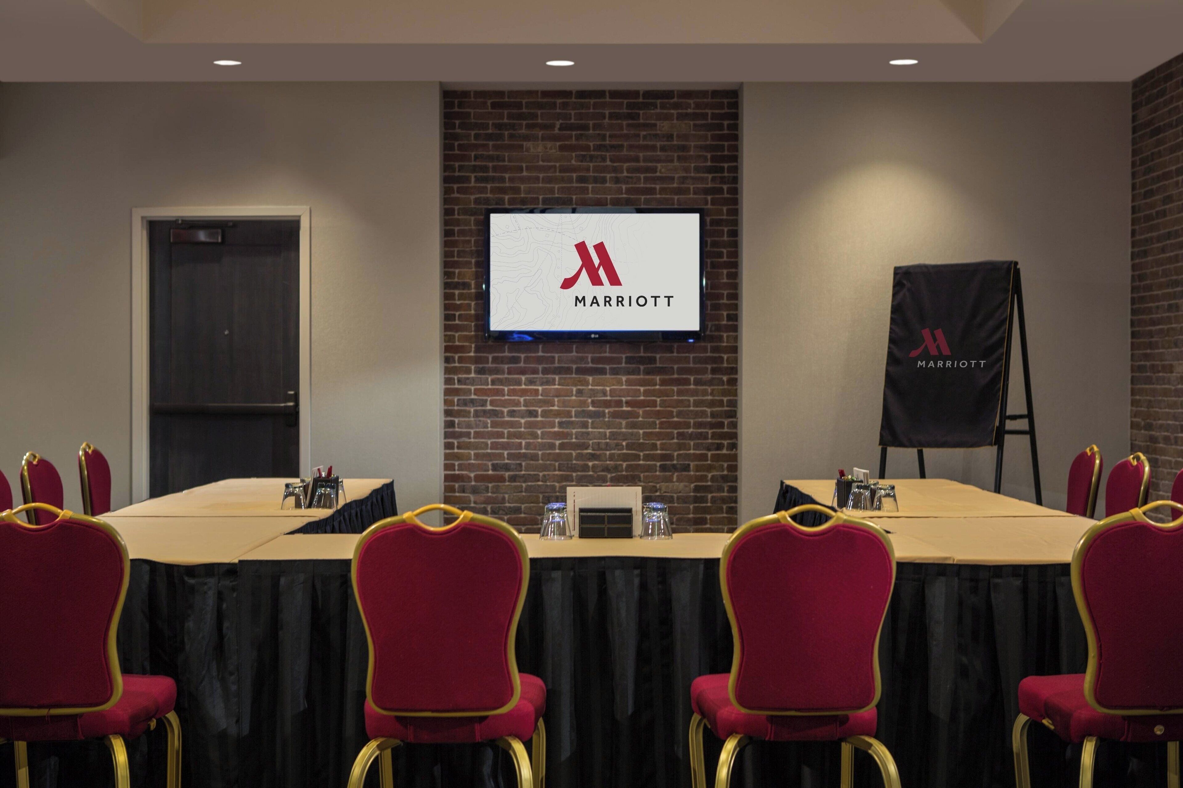 A conference room with a long table, red chairs, a screen displaying the Marriott logo, and a black banner with the Marriott logo.
