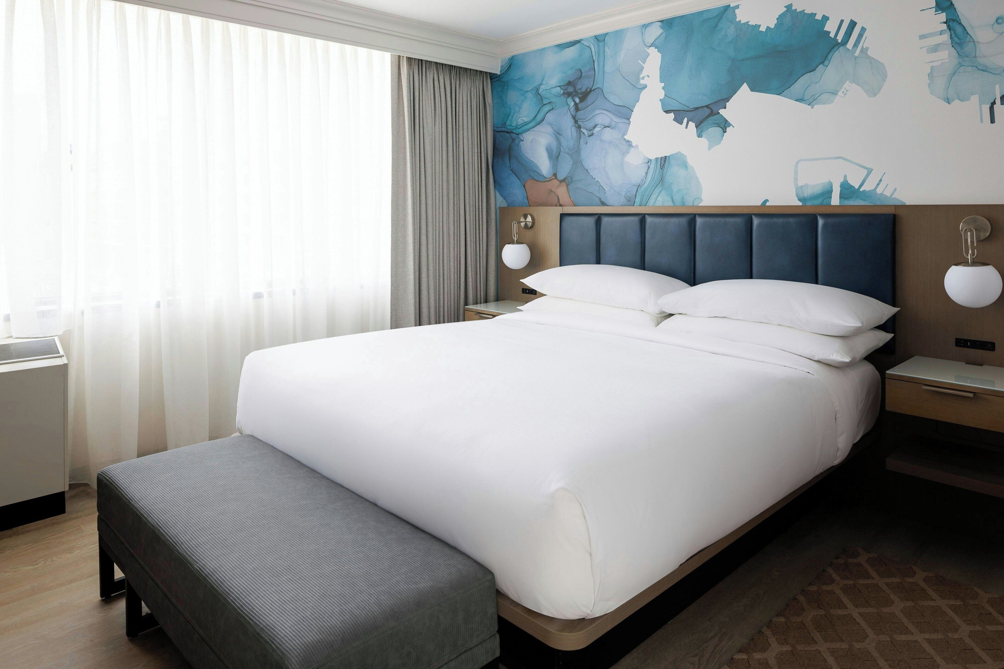 A hotel room with a large bed, a bedside table, a lamp, and a wall with abstract art.