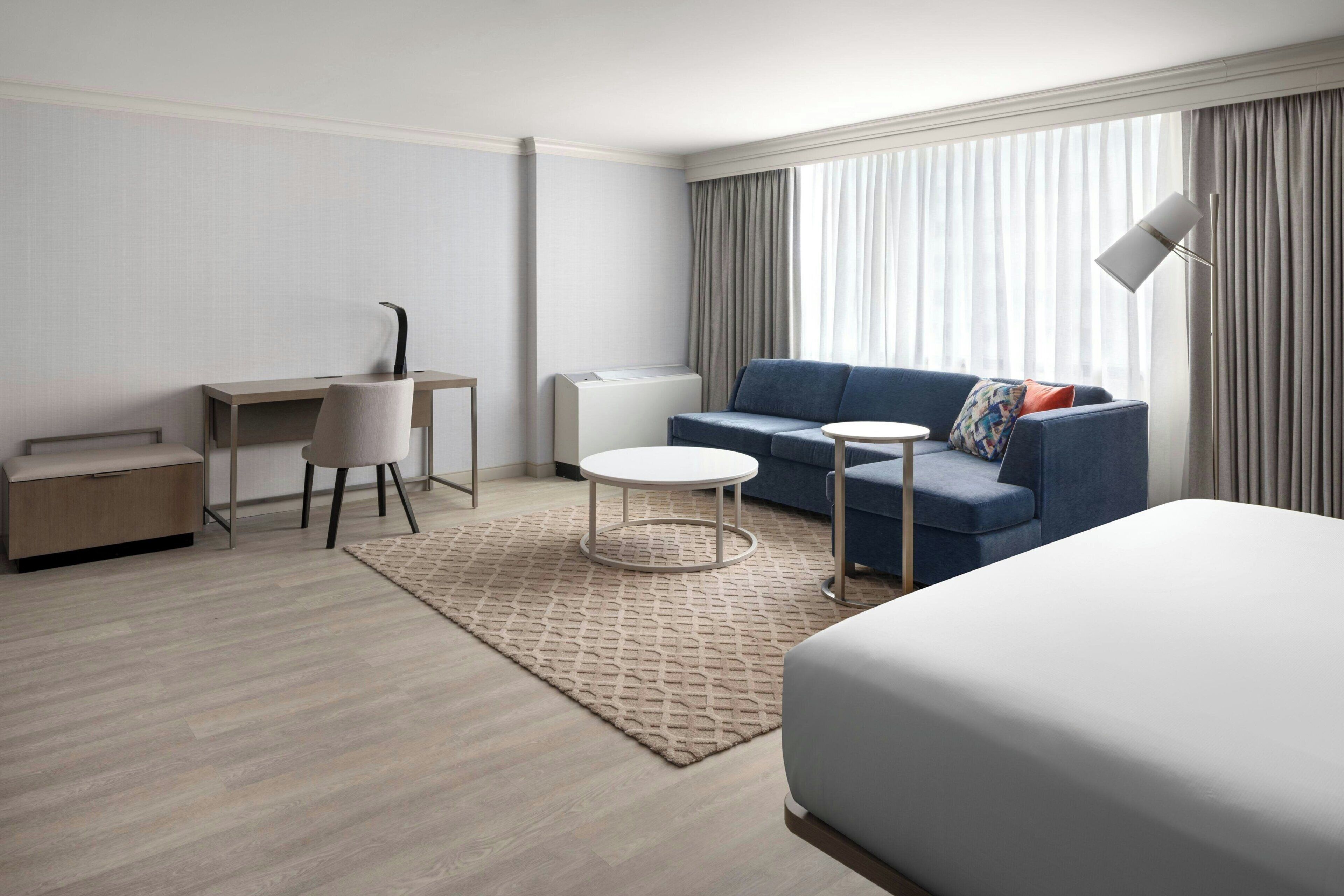 A hotel room with a bed, a desk, a chair, a sofa, a small round table, and a lamp.