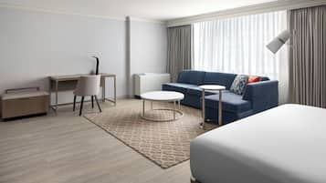 A hotel room with a bed, a desk, a chair, a sofa, a small round table, and a lamp.