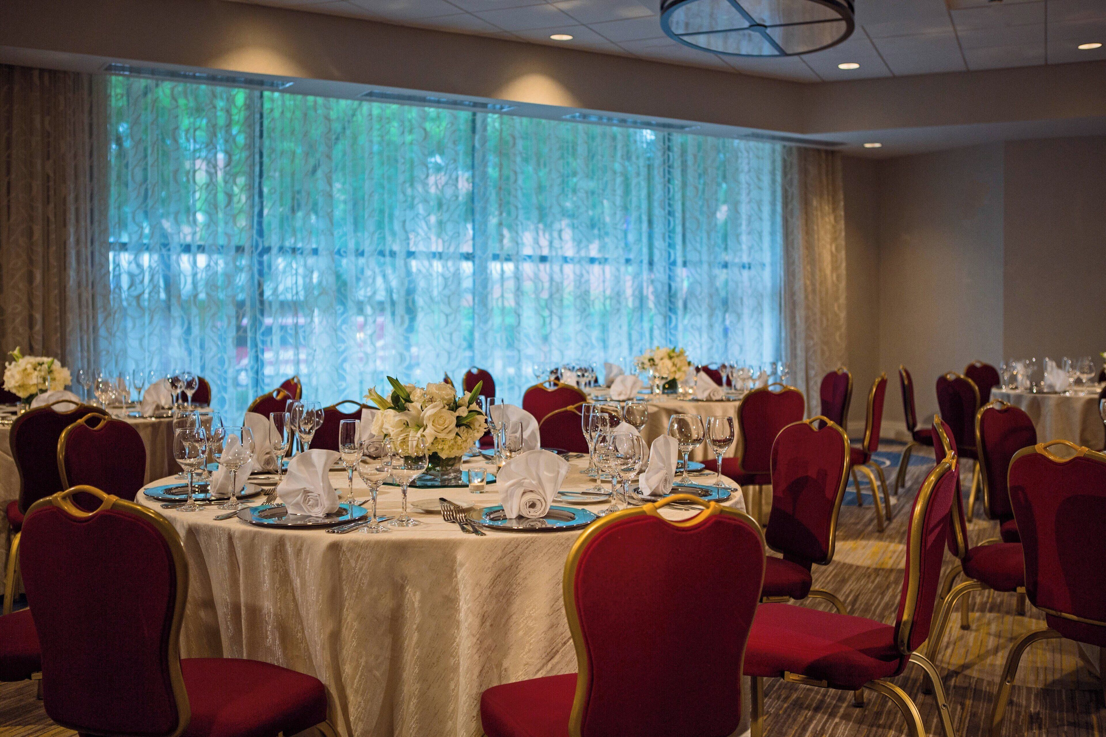 A banquet hall with round tables set for dining, red upholstered chairs, and large windows with sheer curtains.