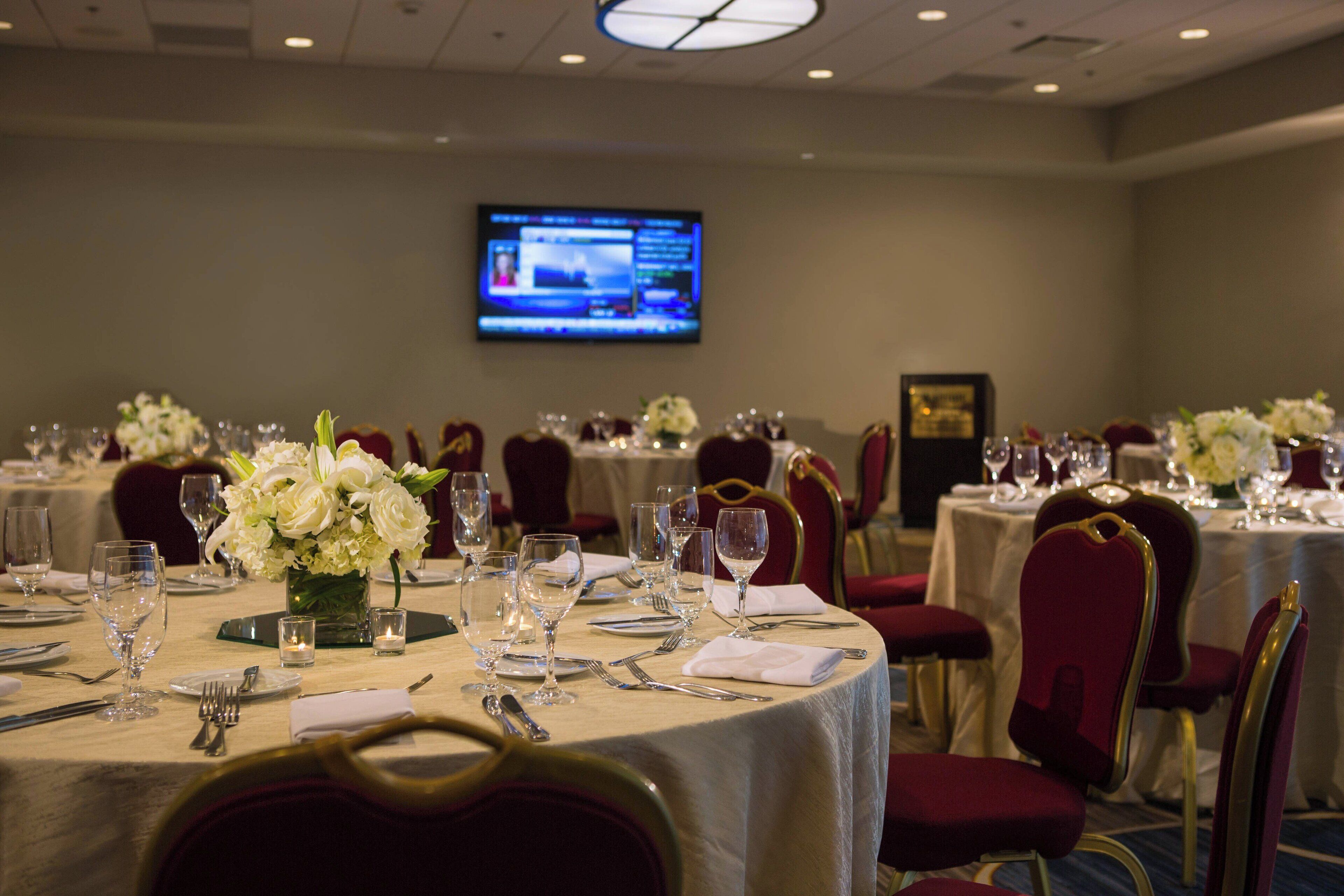 Un salón de conferencias con mesas redondas dispuestas para una reunión, equipado con sillas, cristalería y un centro de mesa floral.