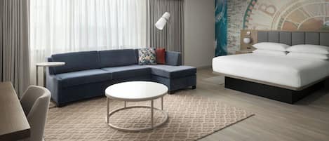 A modern hotel room with a sofa, a round coffee table, a bed, and a large window with curtains.