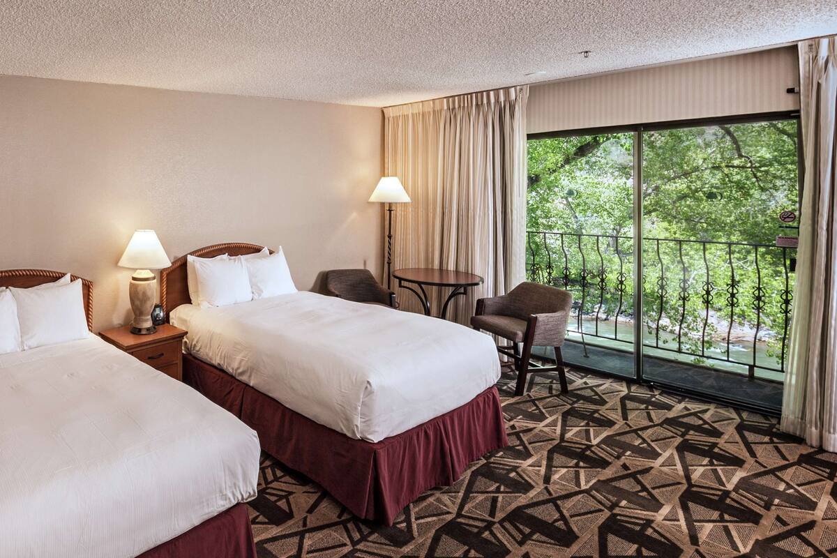 A hotel room with two beds, a small table, a chair, and a balcony with a view of trees.