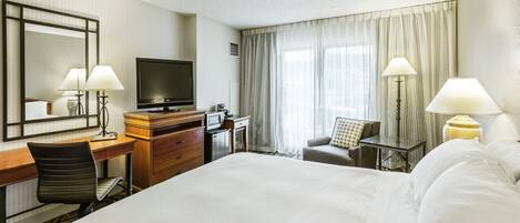 Habitación de hotel con cama, escritorio, silla, televisor y ventana con cortinas.
