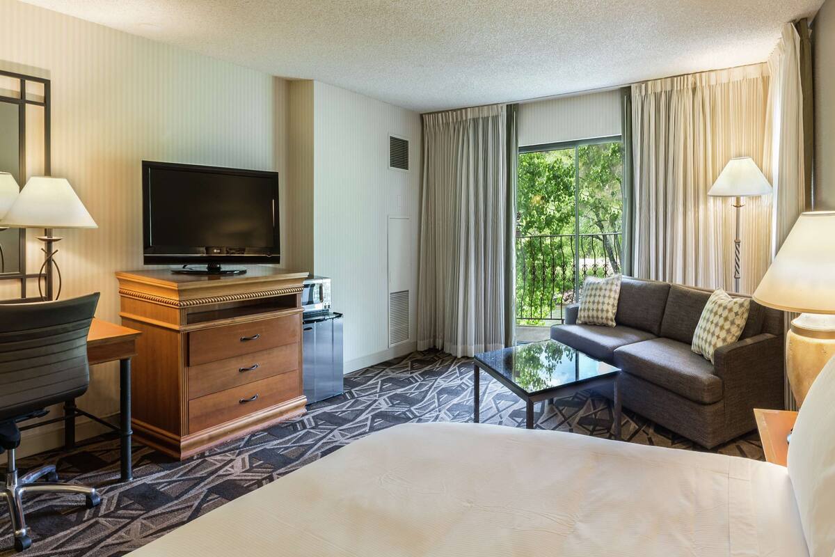 A hotel room with a bed, a desk with a chair, a television, a sofa, and a window with curtains.
