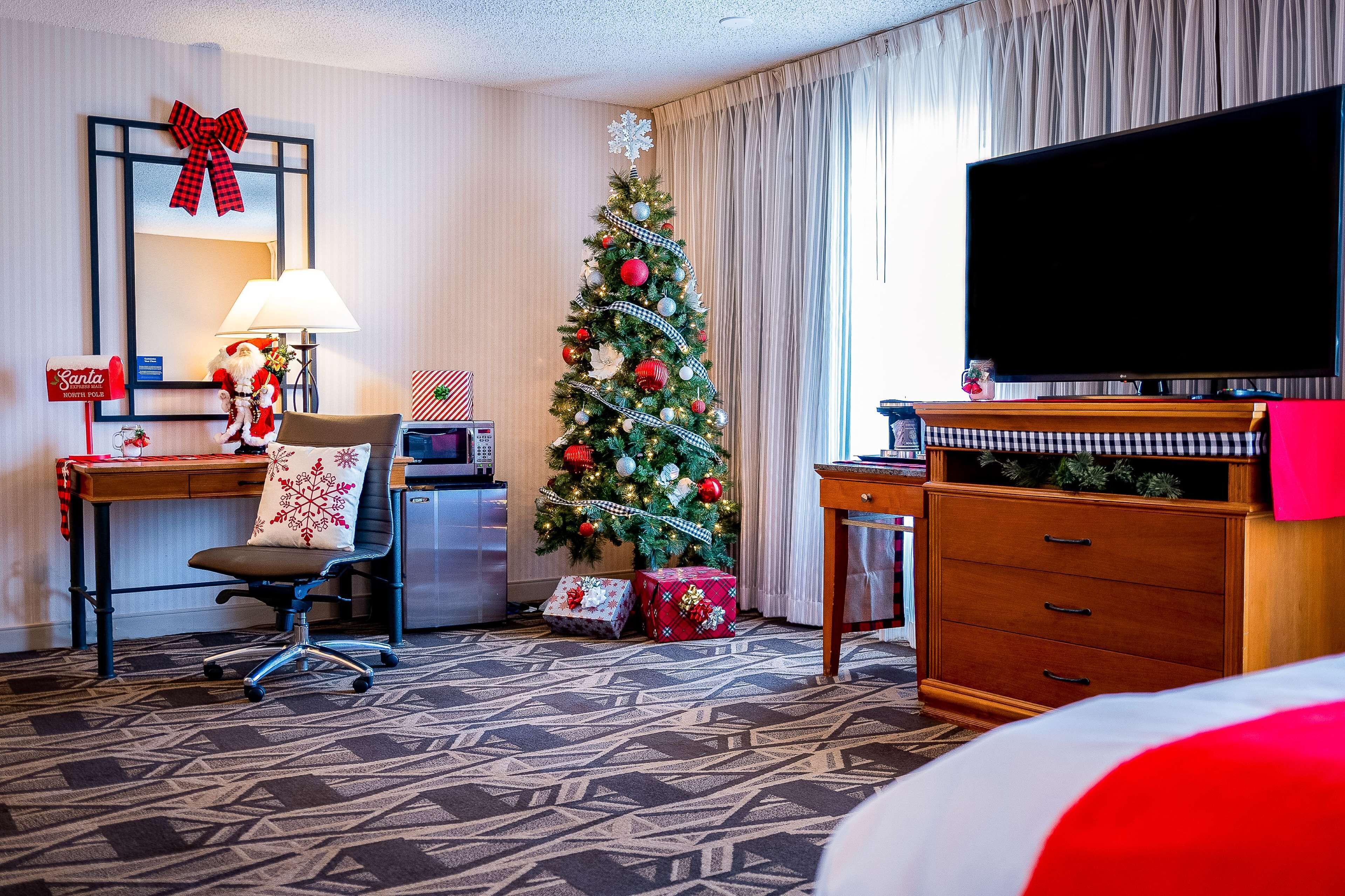 A hotel room with a Christmas tree, a flat-screen TV, a desk with a lamp, and a chair.
