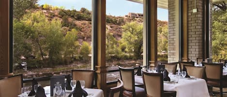 A restaurant with tables set for dining, large windows offering a view of a hillside, and a brick wall.
