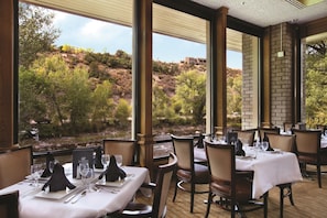 A restaurant with tables set for dining, large windows offering a view of a hillside, and a brick wall.