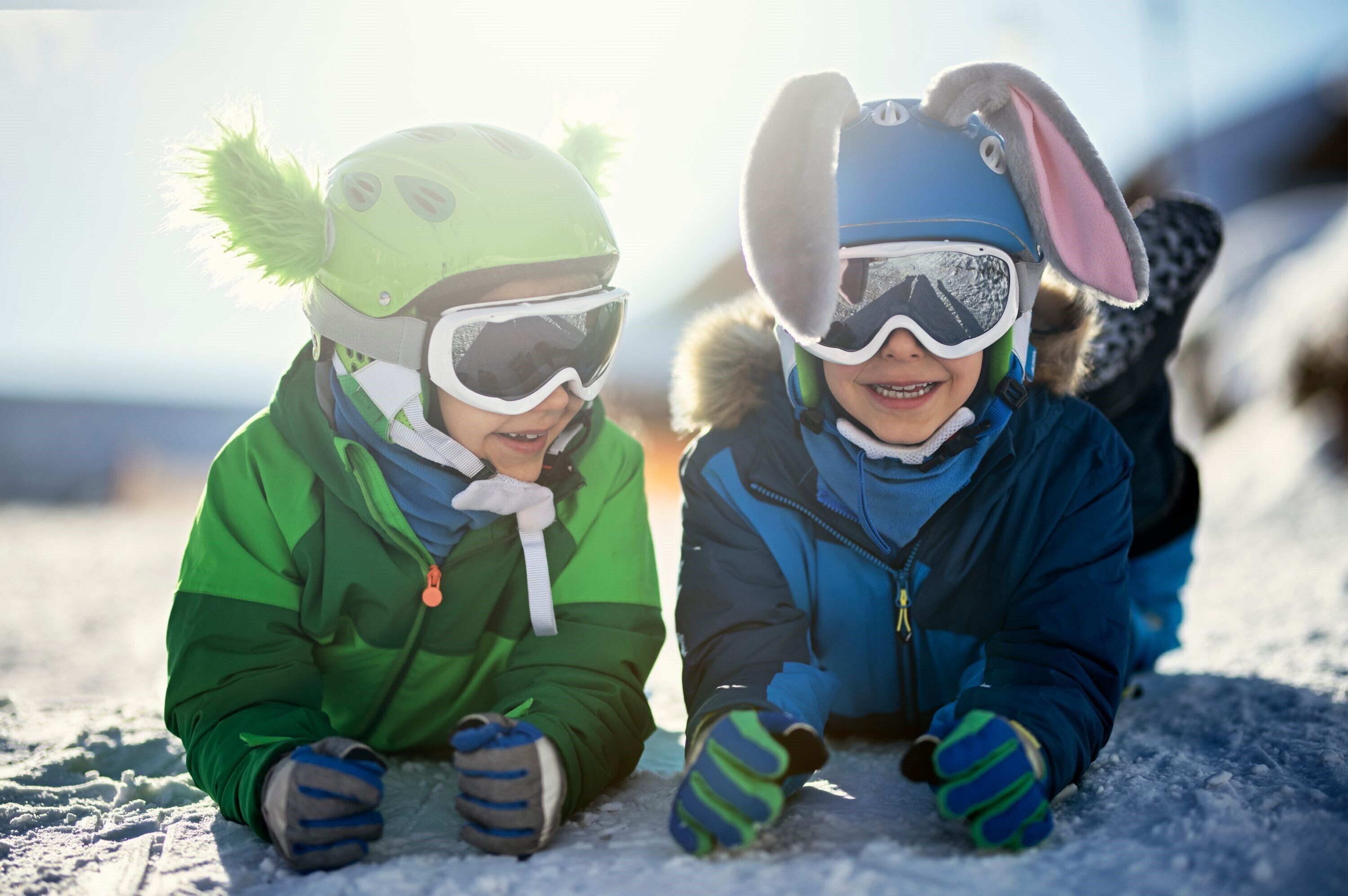 Two children in winter gear and helmets sitting on snow.