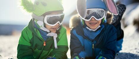 Zwei Kinder mit Winterkleidung und Helmen sitzen im Schnee.
