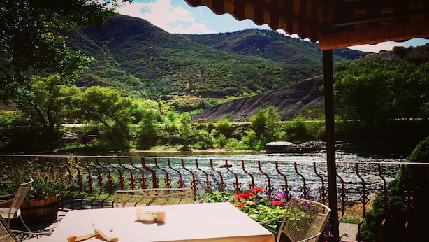 A terrace with a table and chairs overlooking a river, surrounded by greenery and hills.