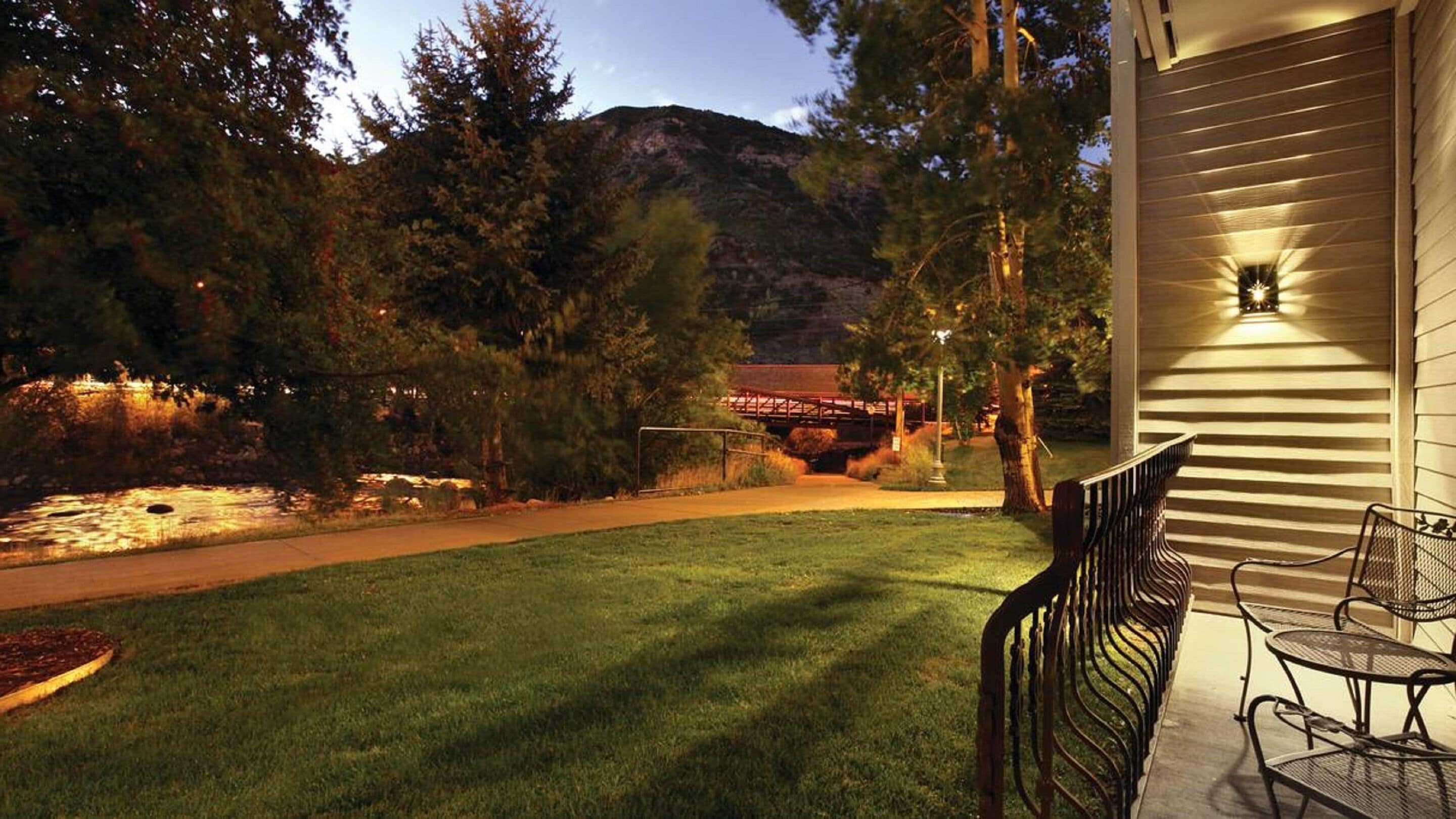 A well-maintained lawn with a metal bench and tables, a pathway leading to a covered area, and a mountain backdrop.