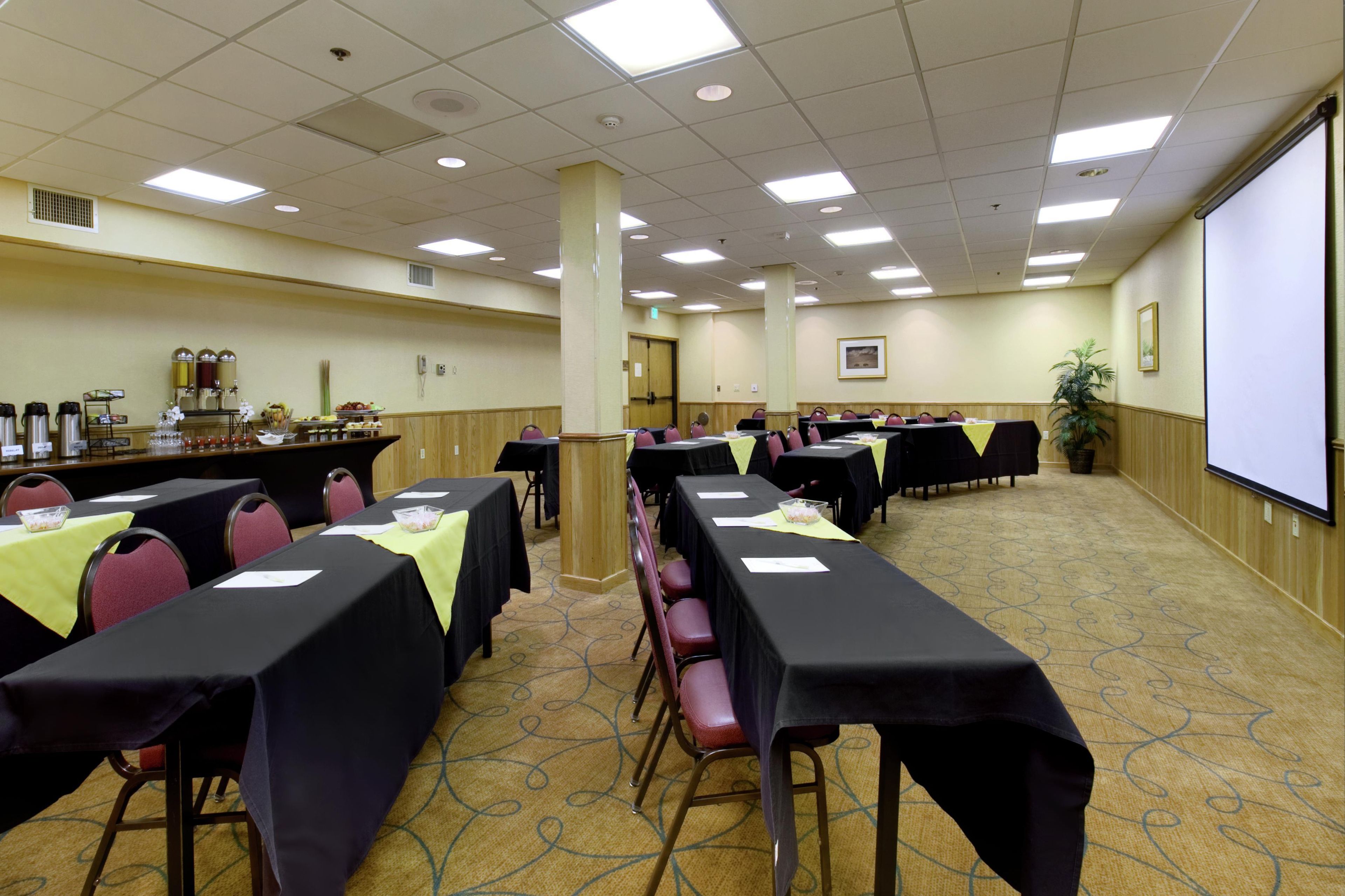 A conference room with rows of tables, chairs, a projector screen, and a buffet area.