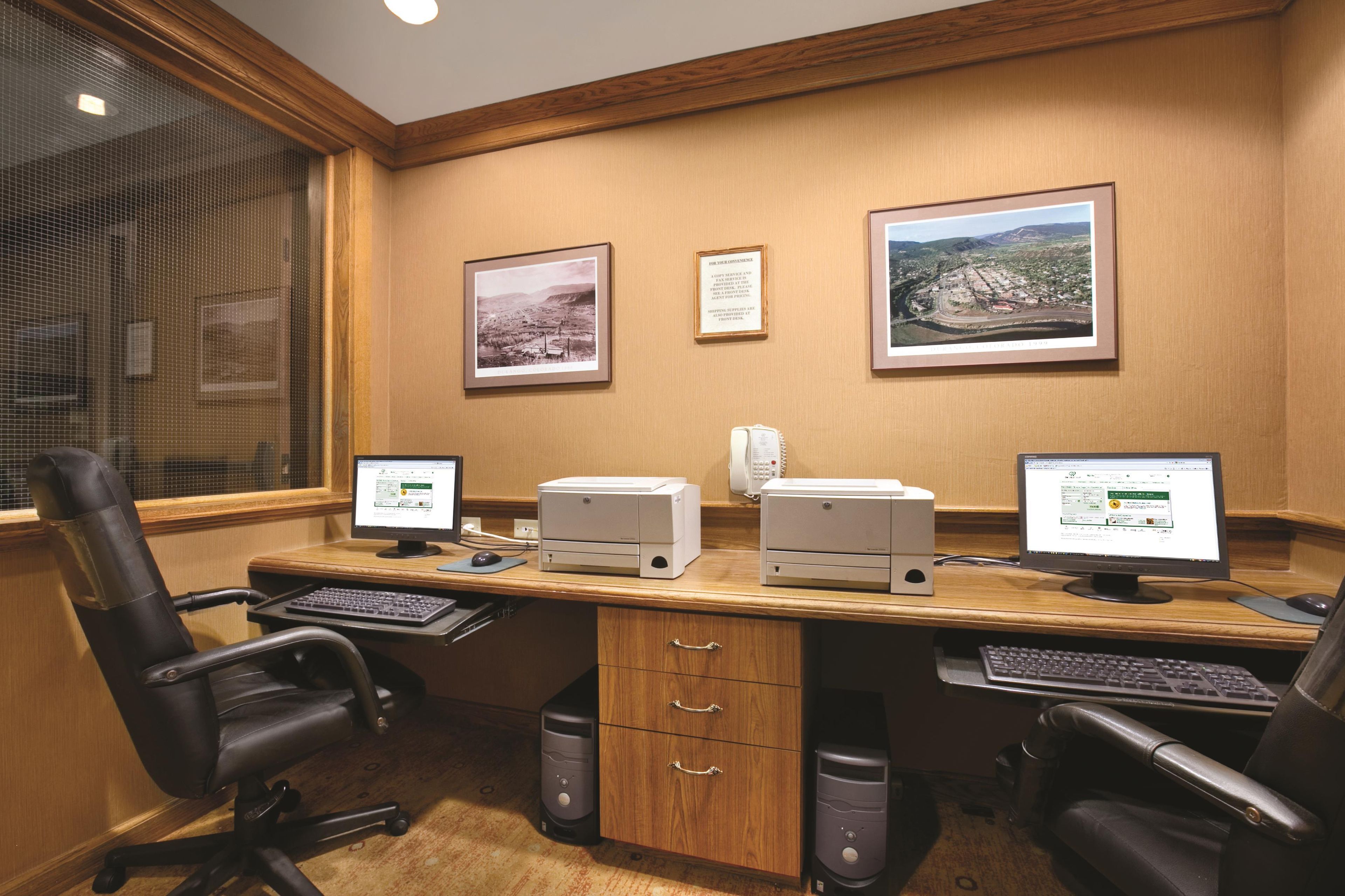 A conference room with four computer stations, two chairs, a wooden desk, and two framed pictures on the wall.