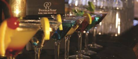 Une rangée de verres à cocktail contenant différentes boissons colorées et décorées de garnitures, un comptoir de bar et, au fond, une boîte étiquetée « Double Tree ».