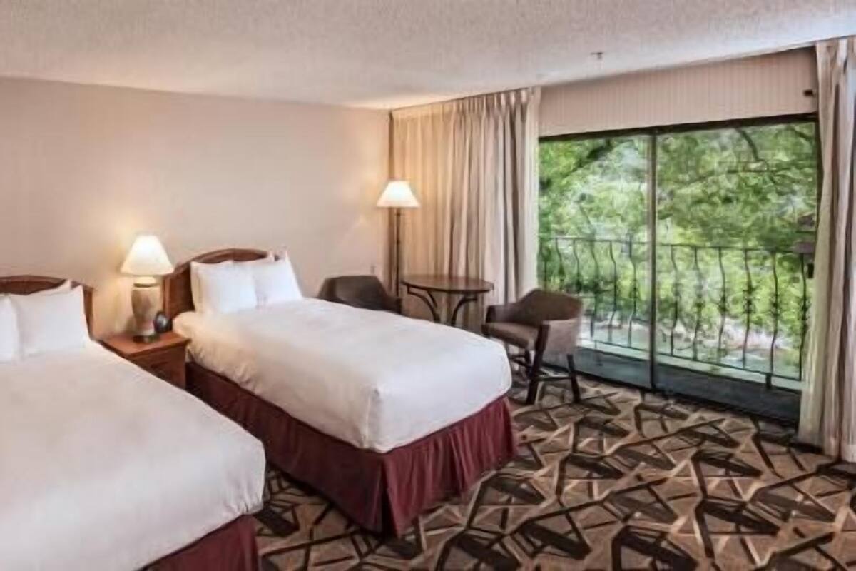 A hotel room with two beds, a small table, a chair, and a balcony with a view of trees.