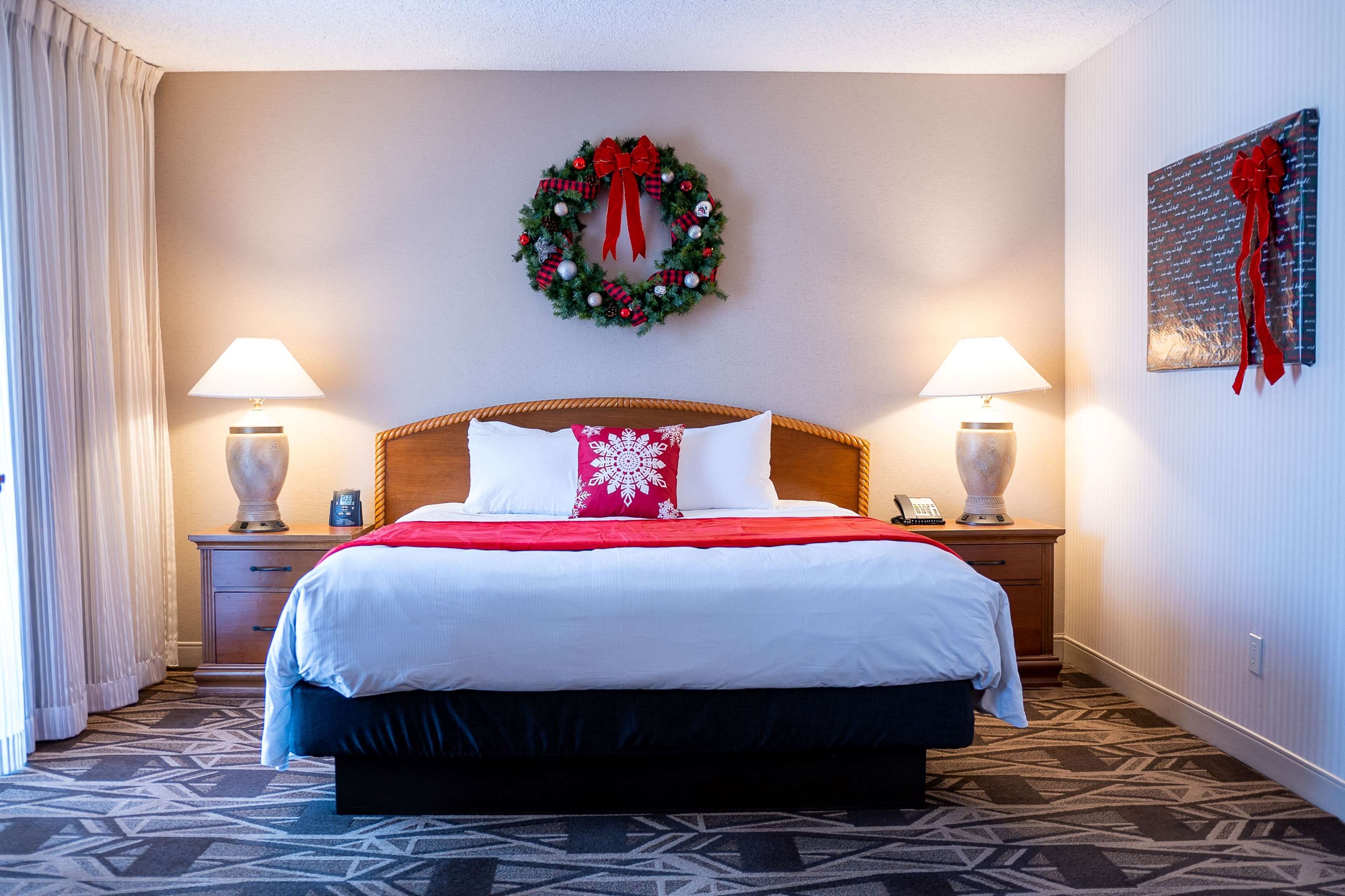 A hotel room with a large bed, two bedside tables with lamps, a red and white pillow, a wall-mounted wreath, and a framed picture with a red ribbon.