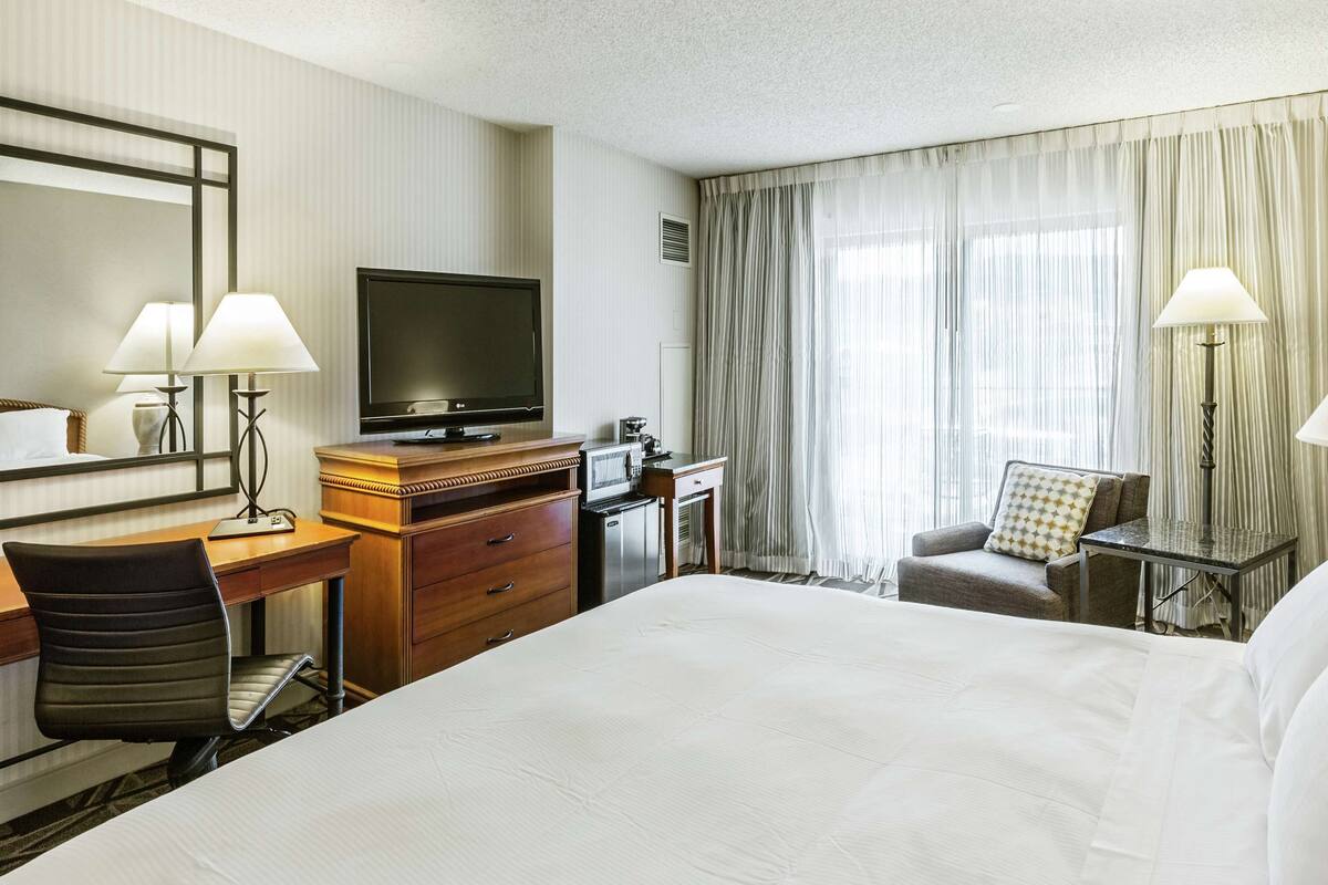 A hotel room with a large bed, a desk with a chair, a television, a minibar, and a window with curtains.