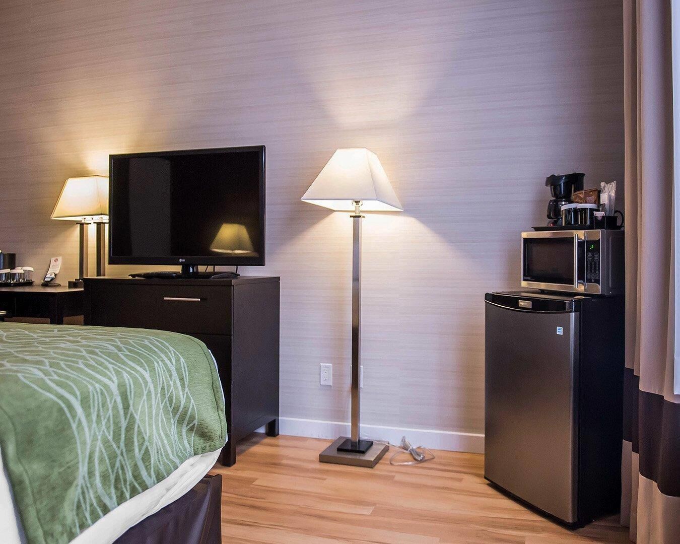 A hotel room with a bed, a desk with a lamp, a television, a microwave, and a refrigerator.