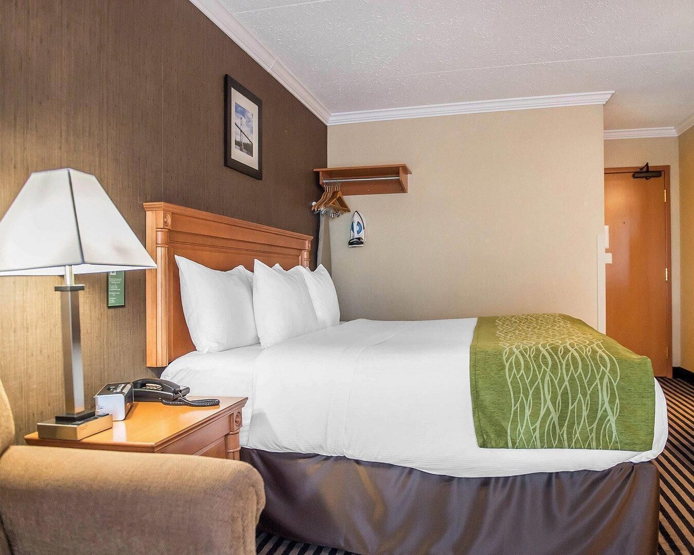 A hotel room with a bed, a bedside table with a lamp, a chair, and a door in the background.