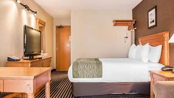 A hotel room with a bed, a desk with a television, a chair, a bedside table, and a wall-mounted lamp.