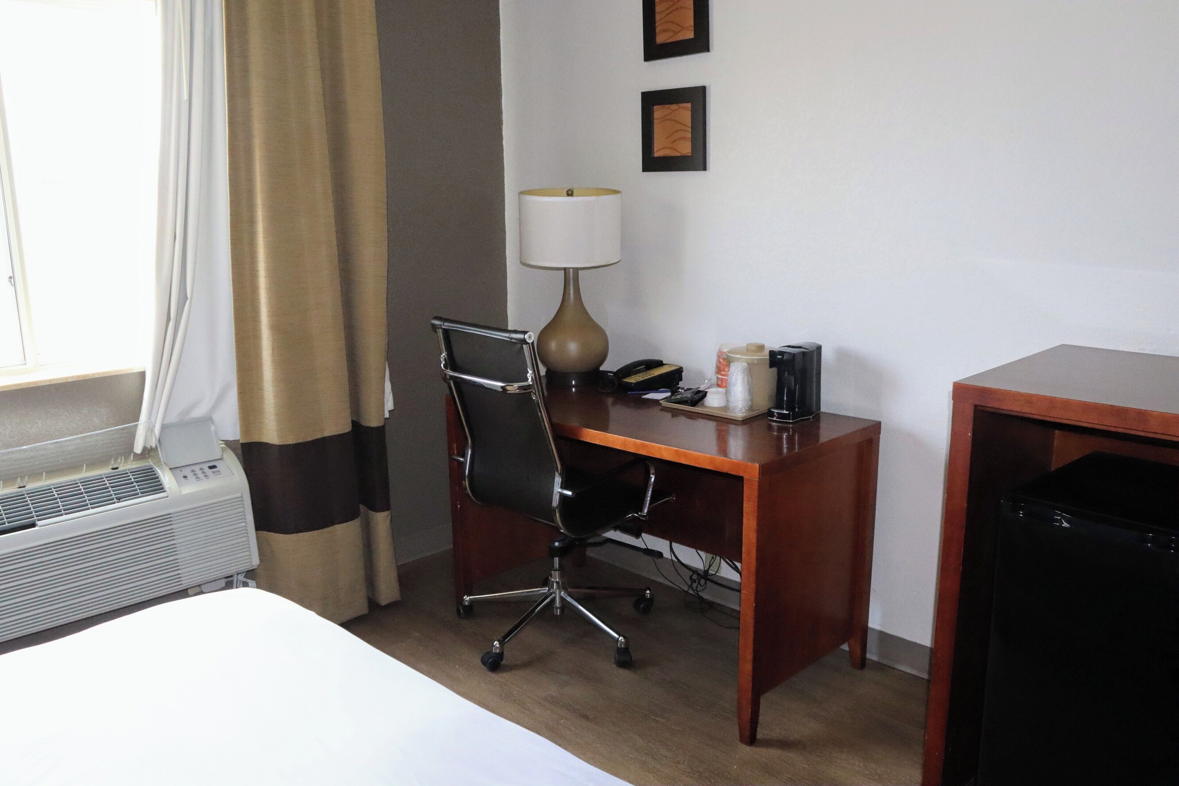 Una habitación de hotel con cama, un escritorio con silla, una lámpara, un teléfono, una mesita y un armario negro.