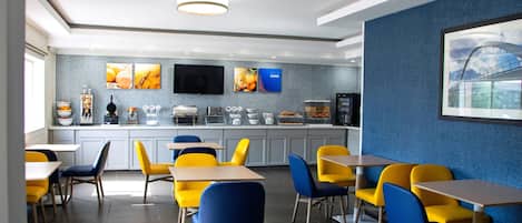 A modern restaurant with yellow and blue chairs, a kitchen area, and a large painting on the wall.