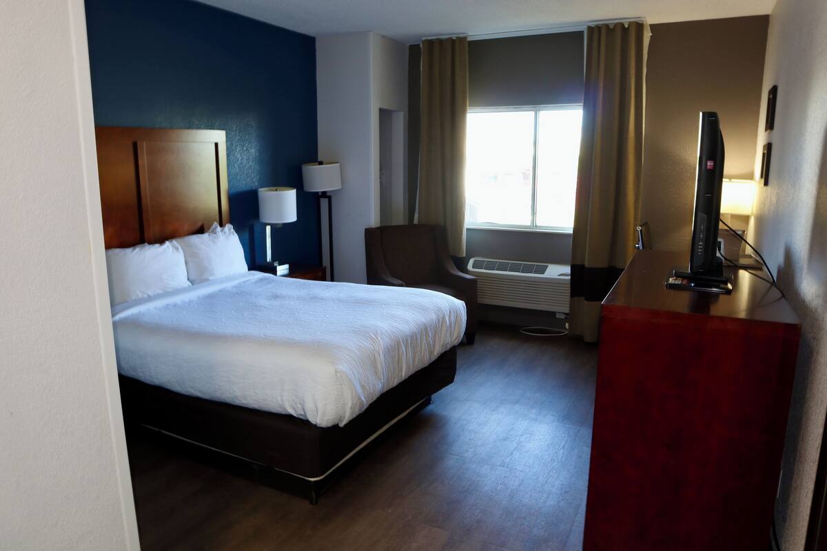 A hotel room with a large bed, a desk with a flat-screen TV, a chair, a lamp, and a window with curtains.