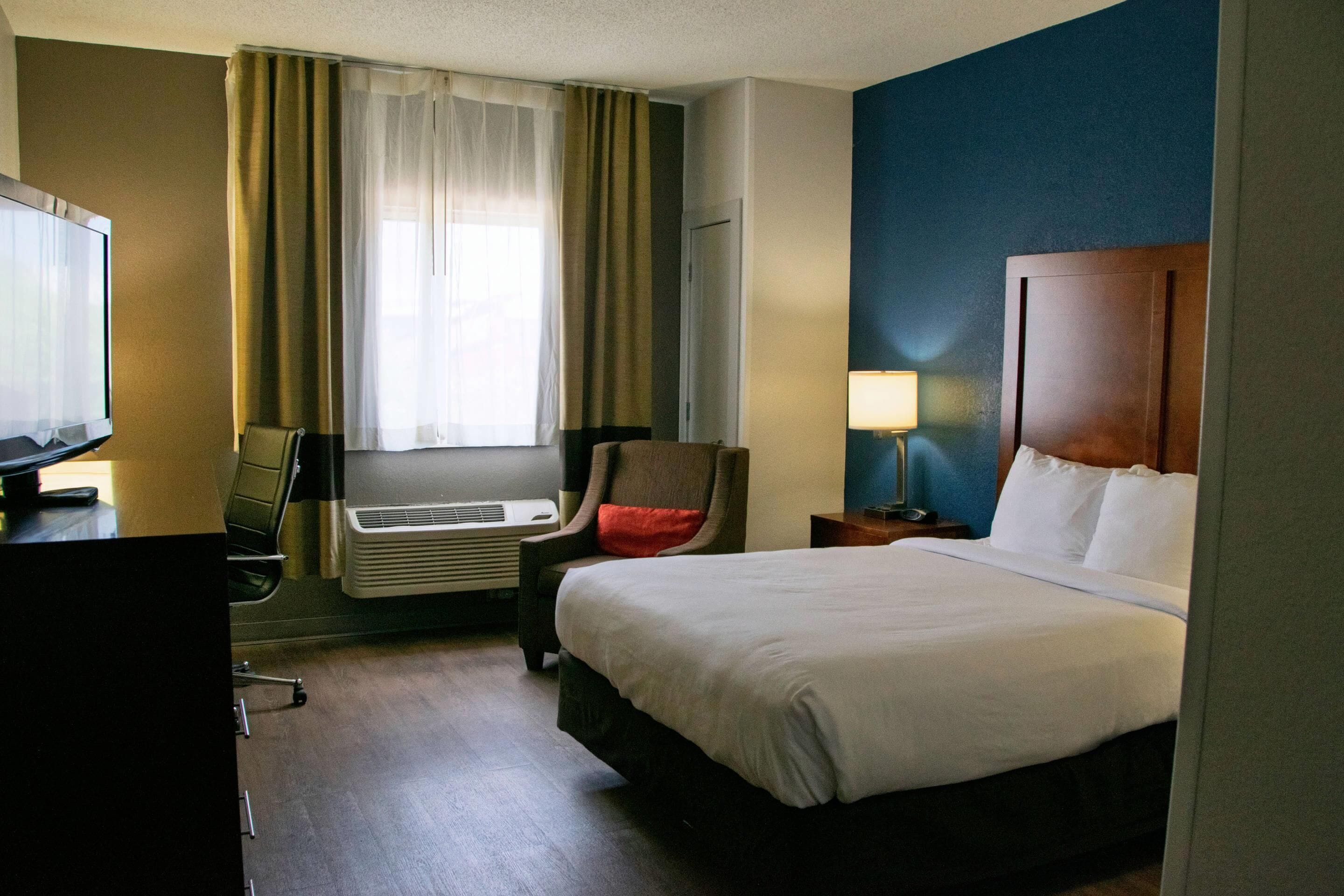 Habitación de hotel con cama, escritorio, computadora, silla, lámpara y ventana con cortinas.