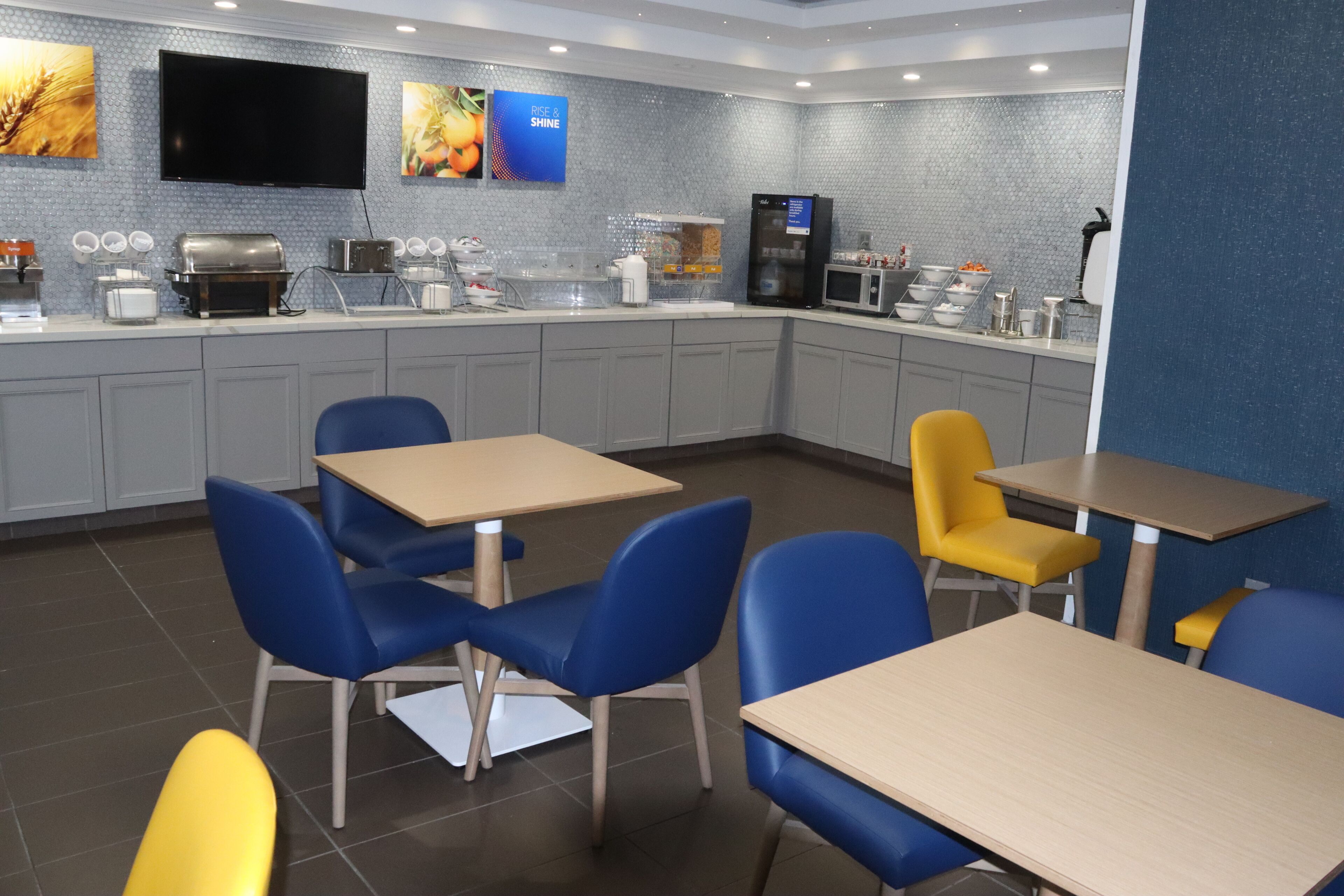 A modern kitchen area with grey cabinets, a microwave, a coffee machine, and a refrigerator. There are blue and yellow chairs around tables, and a flat-screen TV mounted on the wall.