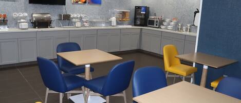A modern kitchen area with grey cabinets, a microwave, a coffee machine, and a refrigerator. There are blue and yellow chairs around tables, and a flat-screen TV mounted on the wall.