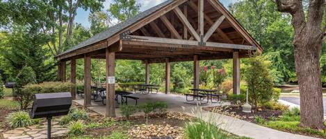 A covered pavilion with picnic tables and a grill area, surrounded by trees and a landscaped garden.