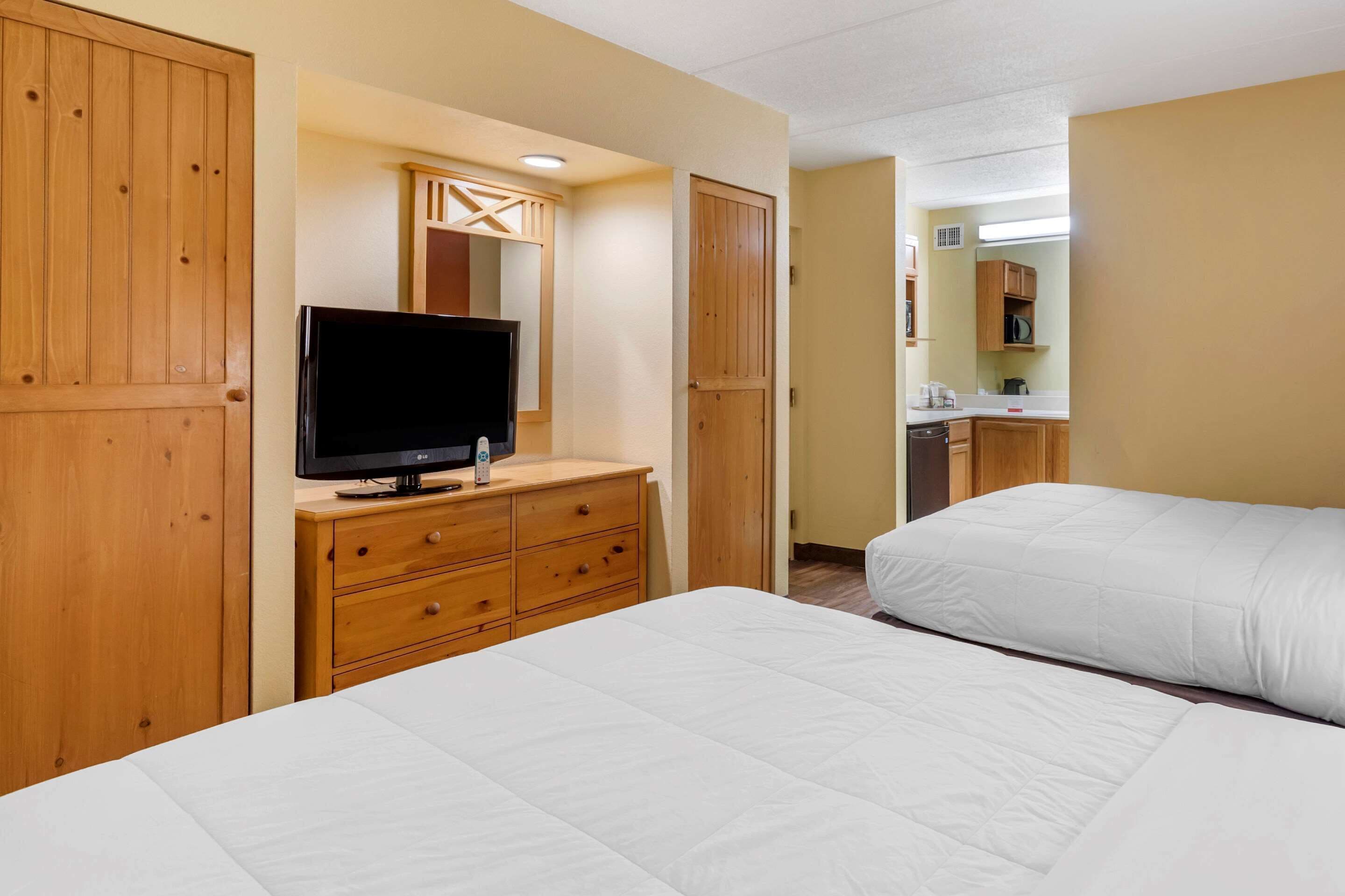 Une chambre d’hôtel avec un lit, un téléviseur posé sur une commode en bois et, au fond, une petite cuisine.