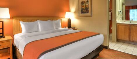 Una habitación de hotel con una cama grande, dos mesitas de noche, un tocador, un baño con espejo y un armario de madera.