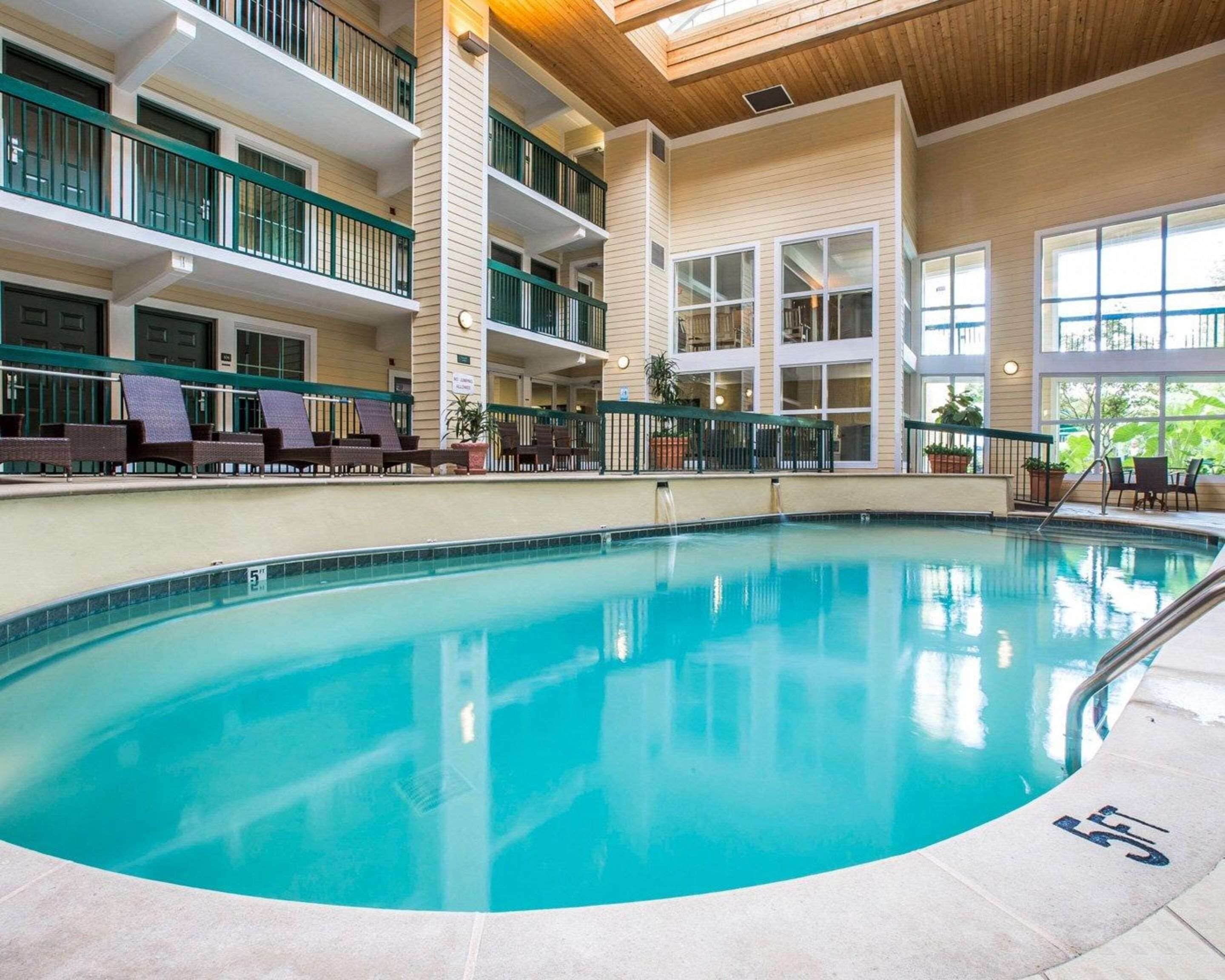 An indoor swimming pool with a clear view of the pool area, lounge chairs, and a multi-level building with balconies.